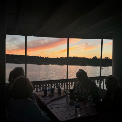 Silhouettes of people sitting at a table with a sunset over a lake in the background.