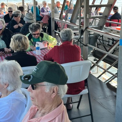 Group of people sitting at tables on a boat with checkered tablecloths and water visible in background.